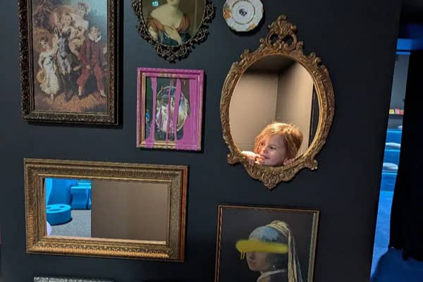 A child's face peeking through an oval frame on a wall of ornate mirrors and framed pictures, including a portrait styled after Girl with a Pearl Earring.