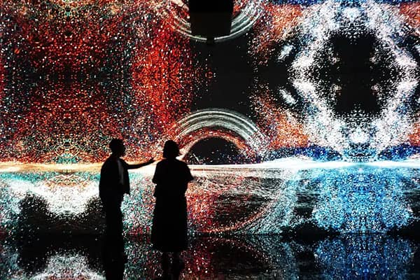 Two silhouetted visitors stand inside an immersive digital art installation, surrounded by colourful red, white and blue light patterns reflected on the floor.