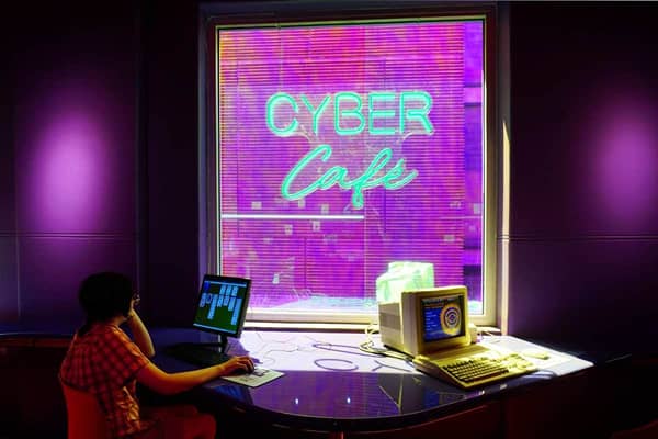 Person in a red checked shirt sitting at a curved desk using a 90s computer, with another beige computer beside them and a large purple-lit window behind showing a green neon sign that reads Cyber Café.
