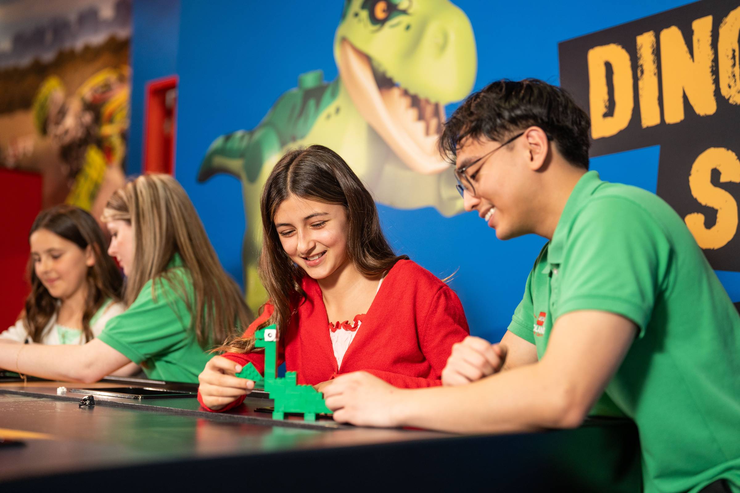 A teenage girl in a red cardigan builds a green Lego dinosaur at a table, guided by a staff member in a green polo shirt, with other guests building alongside them and a large dinosaur mural in the background.