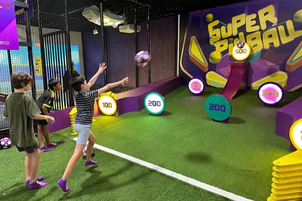Three kids playing Super Pinball, an indoor target game at Superpark Melbourne, with a boy in a striped shirt throwing a ball toward illuminated numbered targets (100 to 500) on an artificial turf court, watched by two other boys.