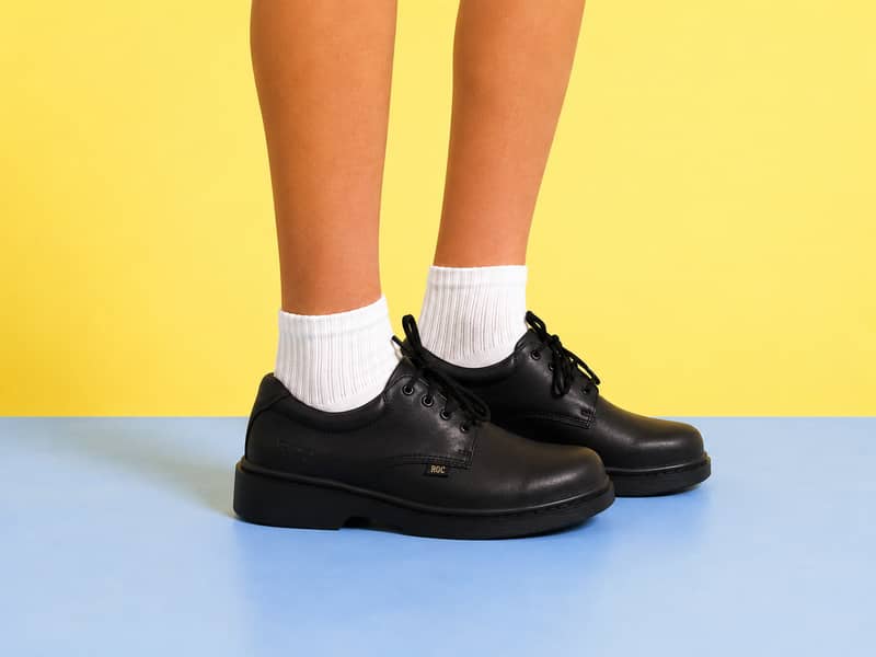 A person's lower legs and feet wearing white ribbed crew socks and low-top black leather lace-up uniform shoes with a small gold "ROC" logo tag on the side. The person is standing against a split background featuring a vibrant yellow top half and a light blue bottom half.