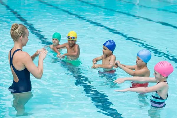 A swim instructor guides five young children wearing colourful swim caps through a group lesson in an indoor pool.