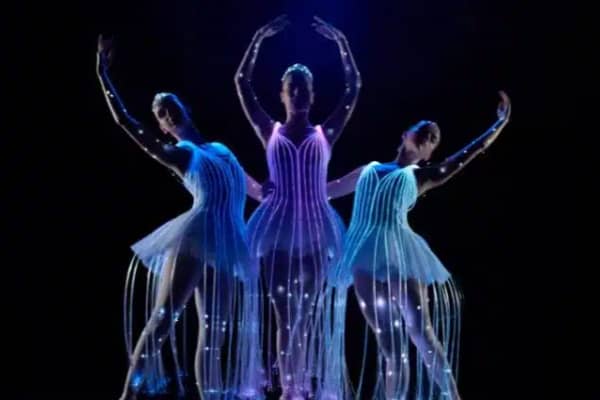 Three ballet dancers perform on a dark stage in illuminated costumes that glow in blue and purple tones, with light trails cascading from their skirts.