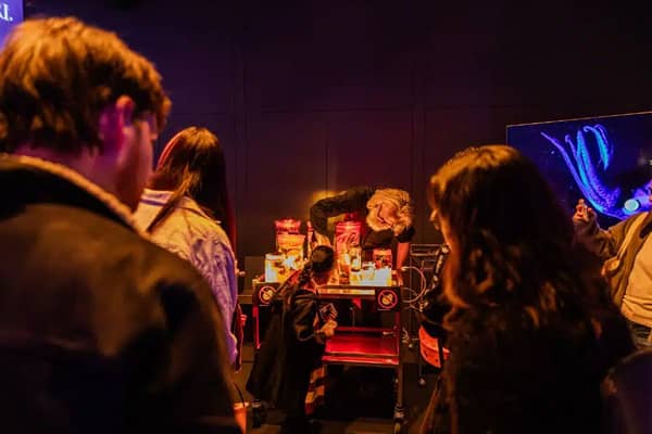A person with grey hair and glasses leans over a small cart of glowing specimen jars in a dim, orange-lit room. A child in a dark costume stands beside the cart, with a crowd of visitors in the foreground and a screen showing a glowing blue sea creature o