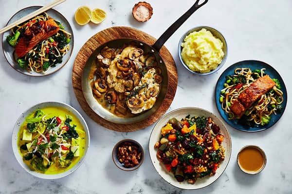 Overhead flat-lay of five meal delivery dishes on a marble surface, including creamy mushroom chicken in a pan, mashed potato, two teriyaki salmon noodle bowls, a green curry bowl and a roasted vegetable and grain salad, with small side bowls of nuts and sauce and chopsticks resting on one dish.