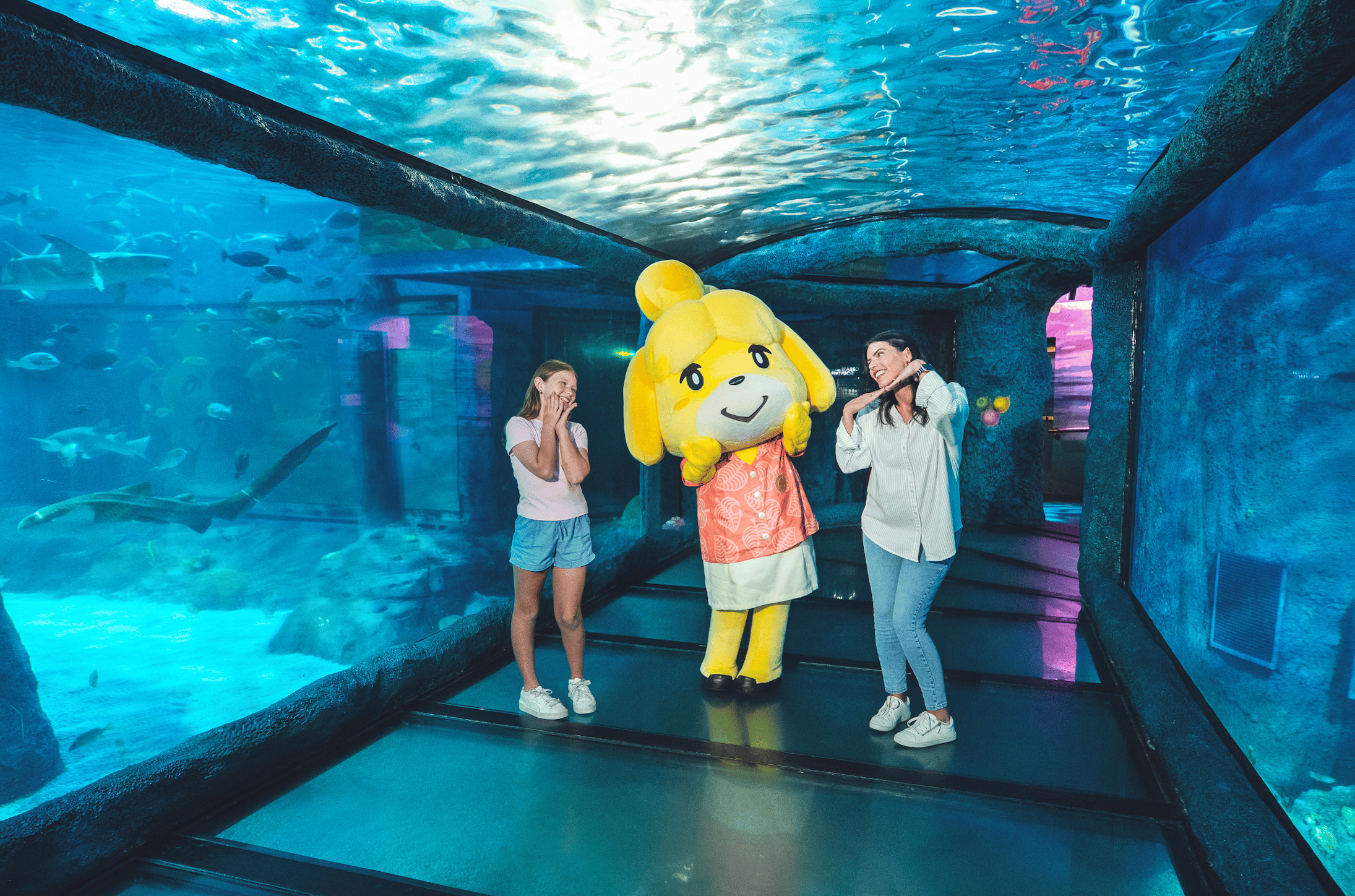 A girl and a woman pose on either side of a yellow dog mascot in an orange leaf-print shirt, inside a glass aquarium tunnel. Sharks and fish swim in the tank on the left, with water overhead.