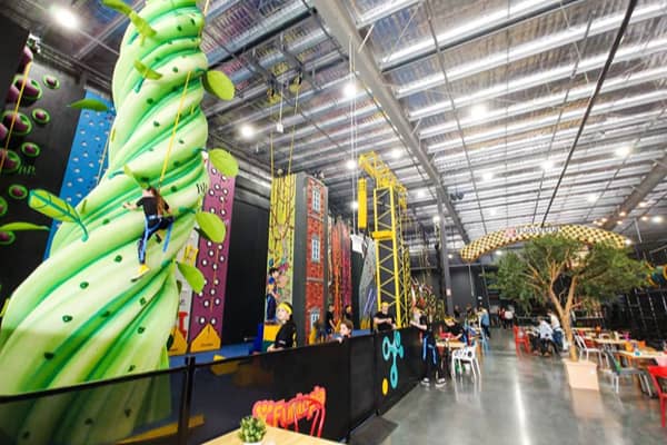 Indoor climbing centre with tall green caterpillar-themed climbing walls and a ropes course, kids climbing among colourful decor, with cafe seating in the background.