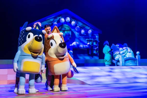 Bluey and Bingo puppets on stage in front of the Heeler family home set during a Bluey's Big Play performance.