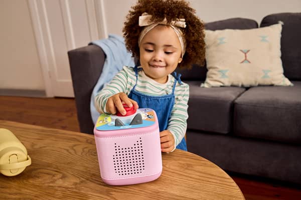 A young girl in a striped top presses a button on a pink Bluey-themed Toniebox 2 speaker sitting on a coffee table at home.