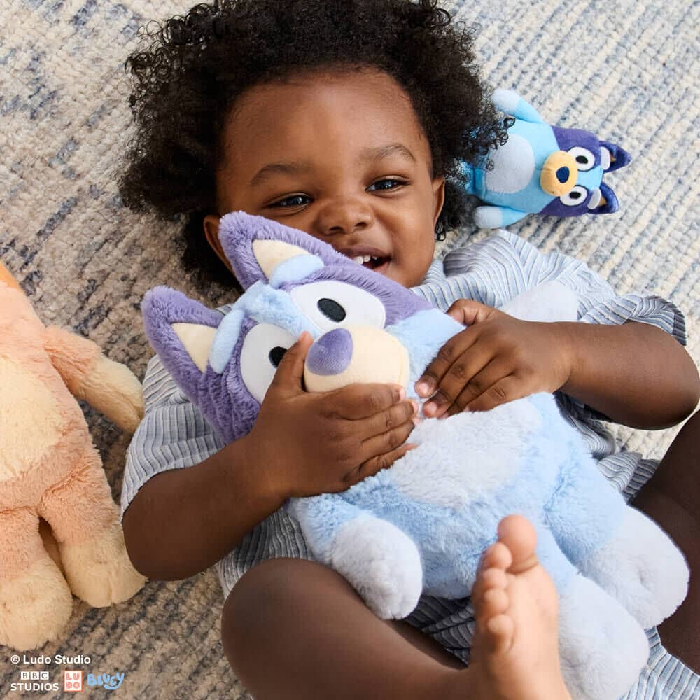 A toddler lying on a rug, smiling and hugging a light blue Bingo plush toy from Bluey, with a purple and blue Muffin plush and a tan plush toy nearby.