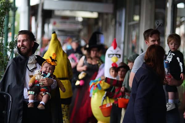A smiling man in a black vampire cape holds a baby in an orange hat with a bat on it, on a shopfront footpath. Behind them, people wear costumes including a banana, a witch hat and a chicken, and a toddler in a skeleton top is held by an adult on the righ