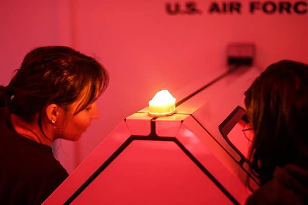 Two people lean in close to examine a small, glowing triangular exhibit display in a dimly lit, red-toned room, with a wall sign reading "U.S. Air Force" visible in the background.