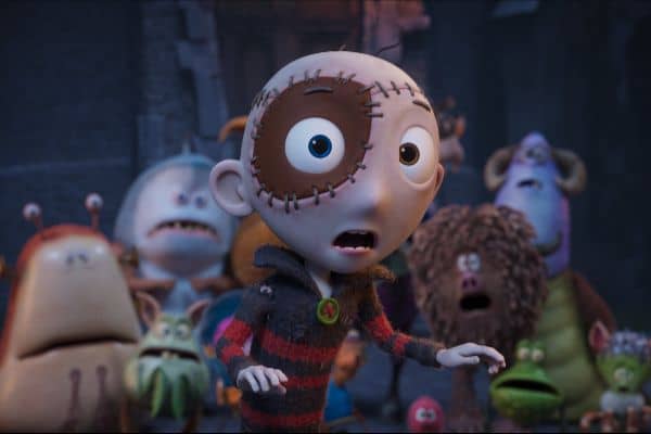 A close-up of the animated character Stitch Head — a small boy with a stitched patchwork face and mismatched eyes — looking surprised, surrounded by a group of monster characters.