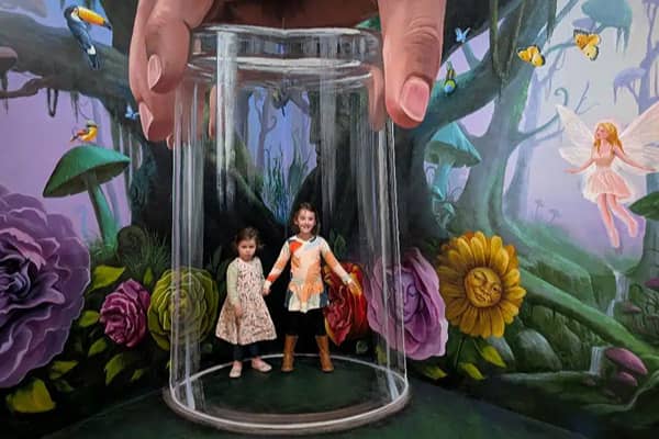 Two young girls stand inside a giant illusion cylinder in an ArtVo mural of an enchanted forest, with a giant hand above them, oversized flowers, a fairy and butterflies