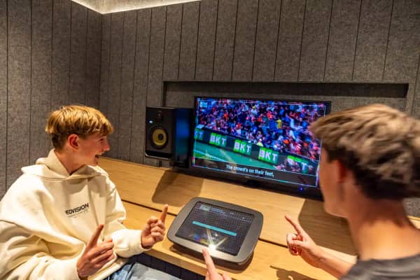 Two teenage boys sit in a commentator's box at the Geelong Sports Museum, calling a football game on the screen in front of them.