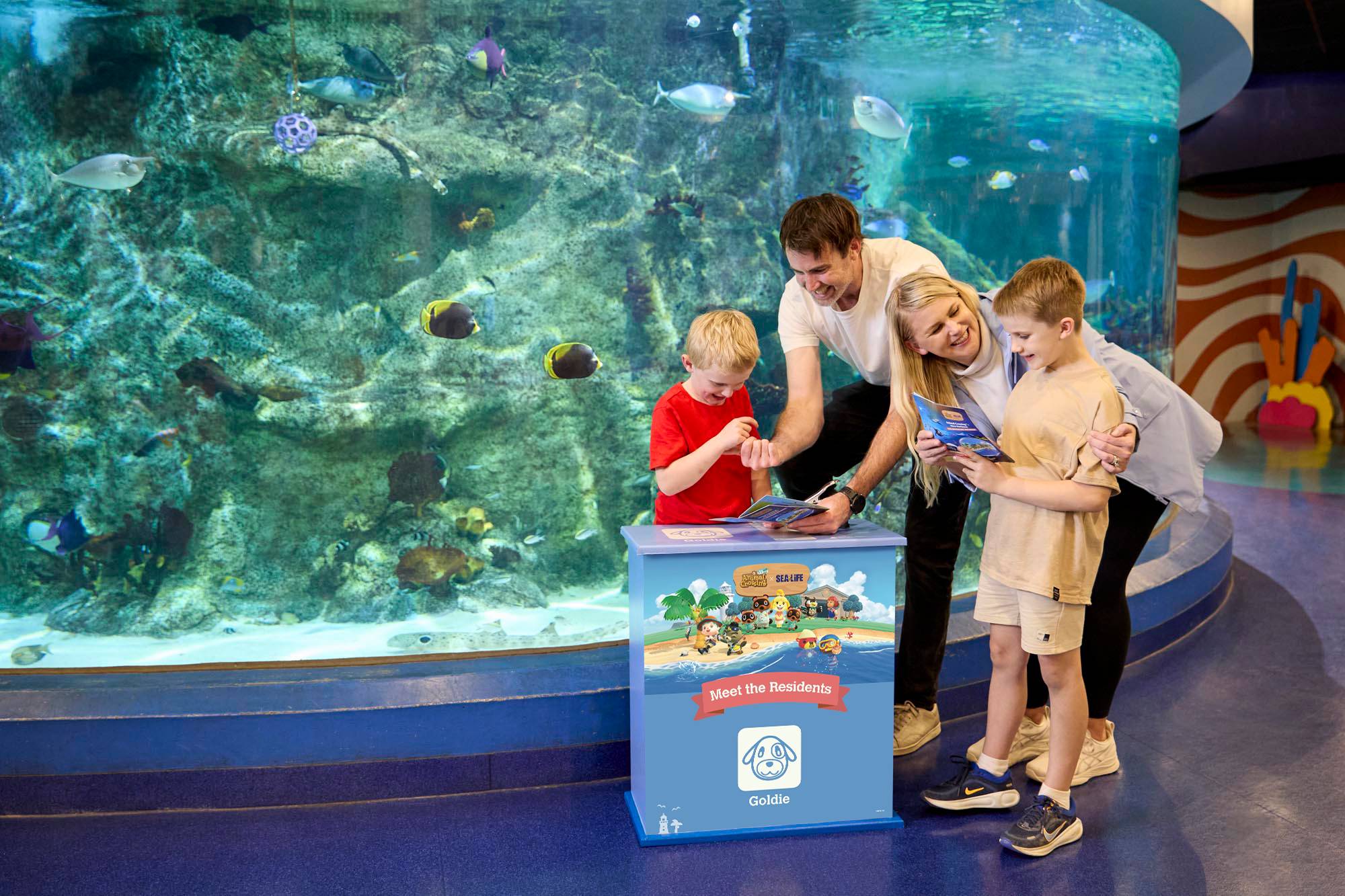 A family of four stands beside a large aquarium tank filled with tropical fish, reading from an Animal Crossing x SEA LIFE branded activity station, a Meet the Residents guide featuring a character named Goldie.