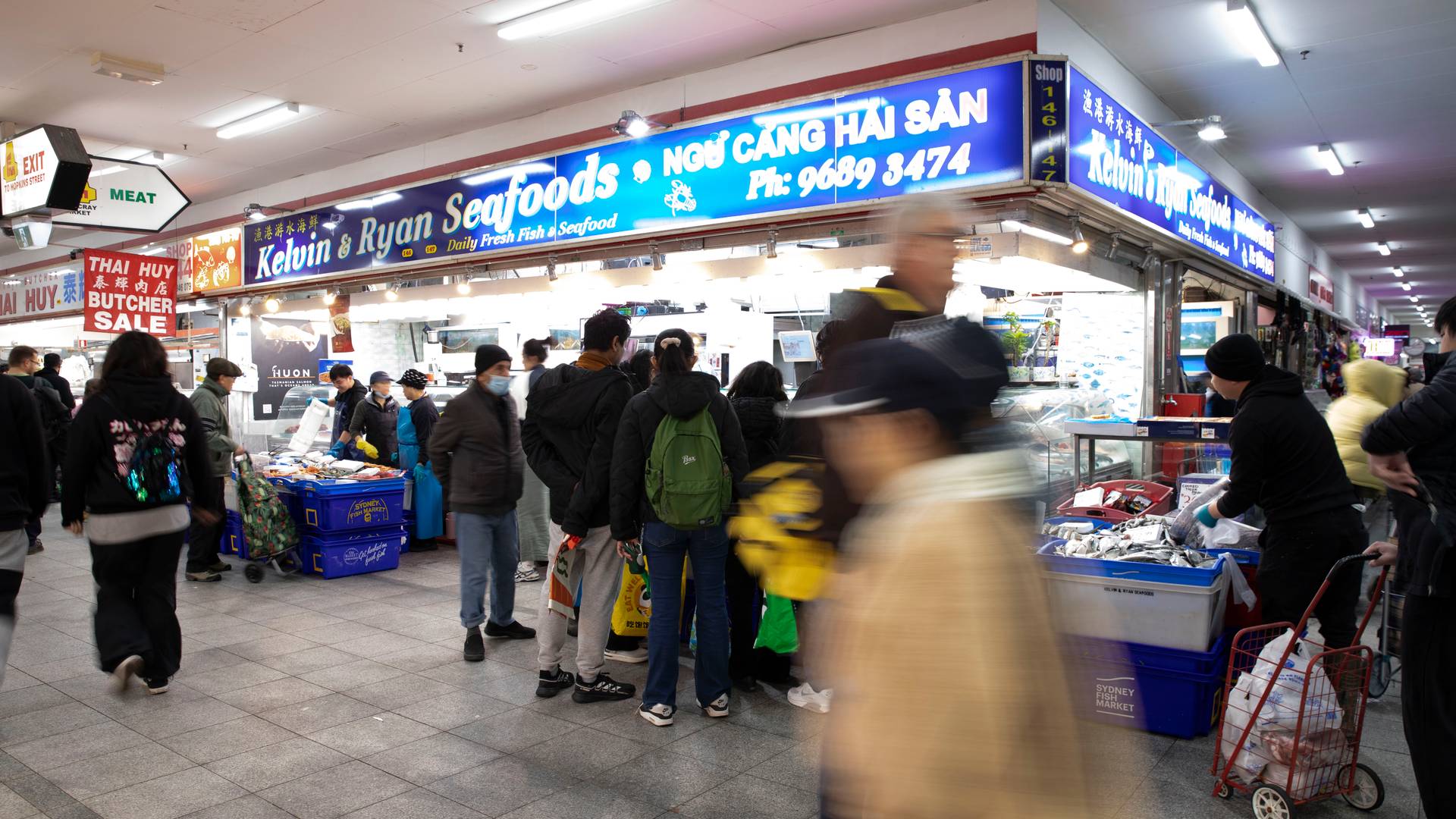 Shoppers, including a young child, queue at a busy seafood stall inside Footscray Market.