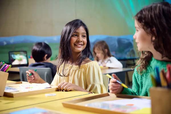 Two children smiling and chatting at a craft table with coloured pencils, paper and drawing supplies at Melbourne Museum's Creative Lab.