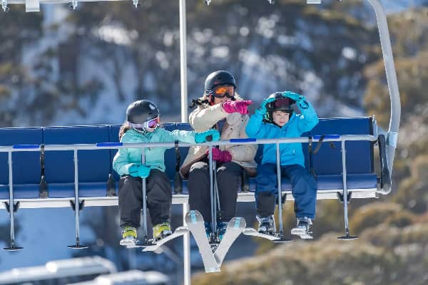 A grown-up and two children in ski gear and helmets ride a chairlift together at Mt Buller on a clear winter day, with snow-covered trees visible in the background.
