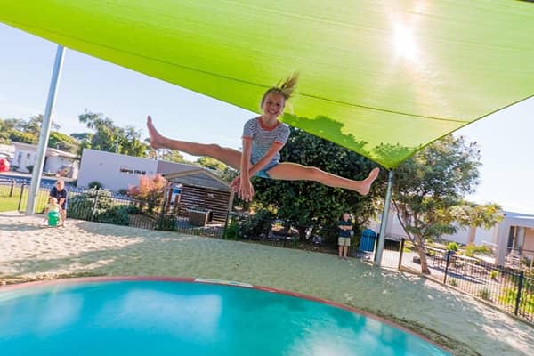 A young girl mid-jump above an in-ground trampoline at an outdoor holiday park play area, legs outstretched, under a green shade sail. Sand surrounds the trampoline, with low fencing, cabins and other children playing in the background on a sunny day.