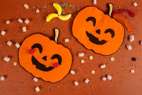 Two flat orange pumpkin-shaped treats with smiling black faces, one with red lolly cheeks, lying on an orange background. They are surrounded by rainbow sprinkles, mini marshmallows, white buttons and gummy lollies.