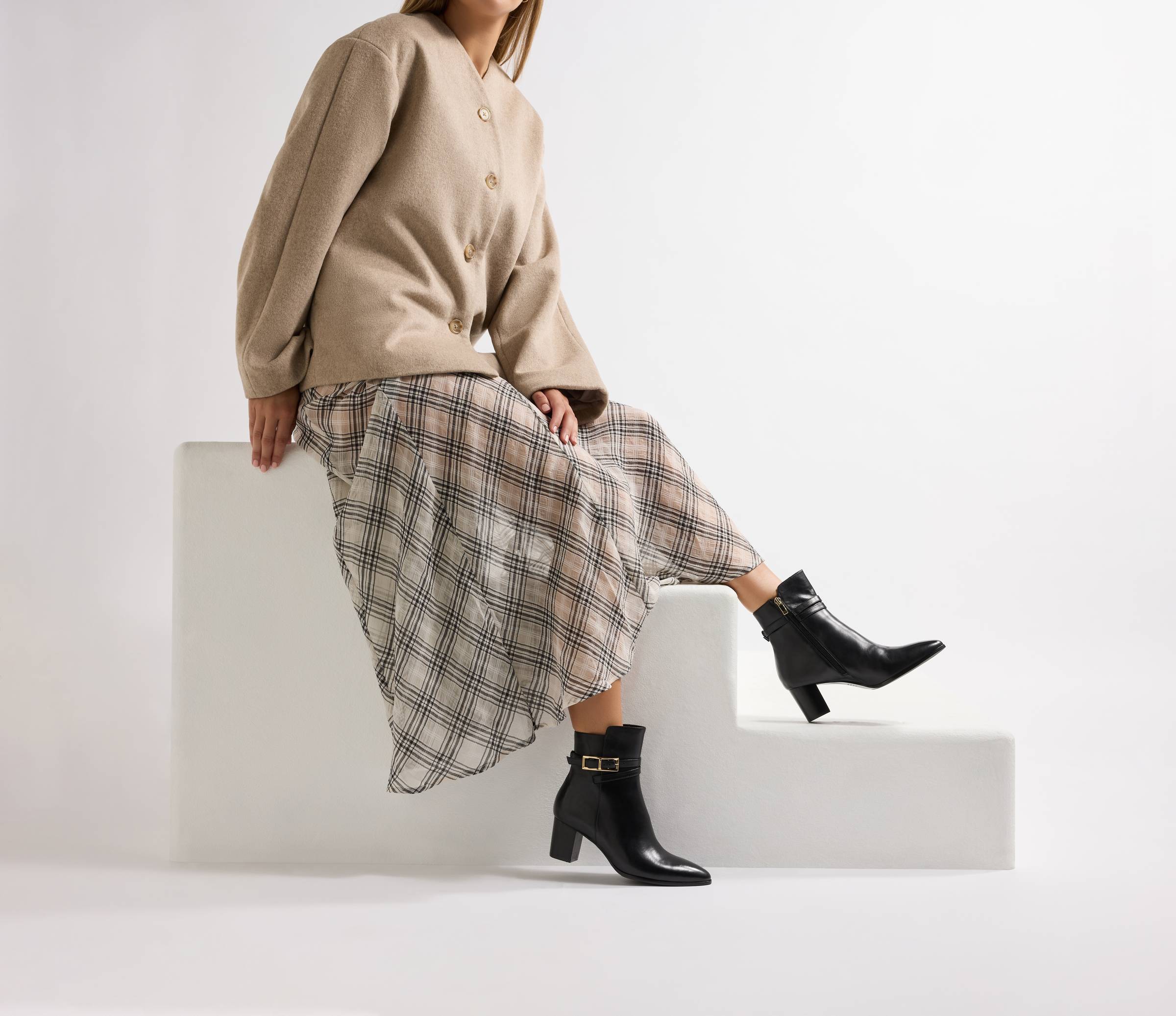 A person sits on a white stepped block wearing black heeled ankle boots with a gold buckle detail, styled with a plaid midi skirt and a camel cardigan.
