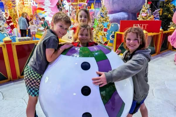 Four happy children gather around a giant oversized Christmas bauble decoration, smiling at the camera against a backdrop of colourful festive displays, fairy lights, Christmas trees and character installations at Melbourne's Christmas Wonderland.