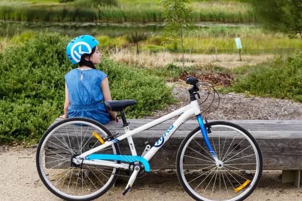 A child wearing a blue helmet sits on a wooden bench beside a white and blue Byk bike, looking out over native bushland.