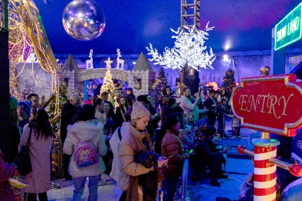Families explore a festive indoor winter wonderland under a big top, with Christmas trees, glittering decorations, a large mirror ball, and a Snow Land sign visible in the background.