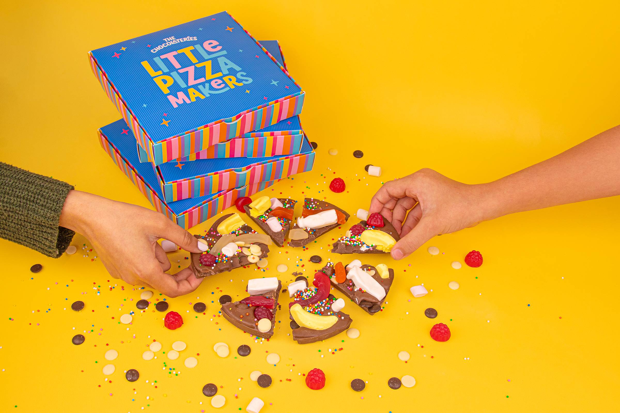 Two people's hands reaching in to take slices of chocolate pizza topped with lollies, marshmallows, sprinkles and chocolate buttons, arranged in a circle on a yellow background. A stack of striped Little Pizza Makers boxes from The Chocolateries sits behind, with loose lollies and sprinkles scattered across the surface.