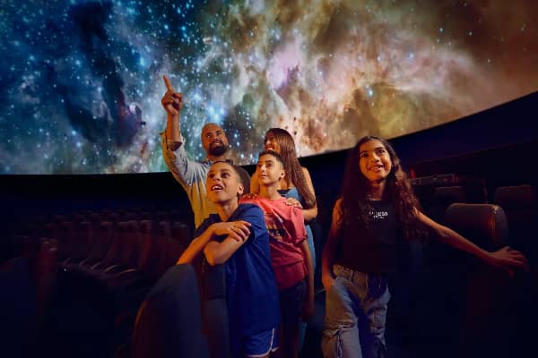 A parent and four kids look up in wonder at a sweeping space projection covering the ceiling of a large dome theatre.