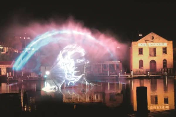 A dramatic outdoor light show at Flagstaff Hill Maritime Village in Warrnambool, with a large face projected onto a mist screen over the water, surrounded by sweeping beams of pink and blue light reflecting off the harbour at night.