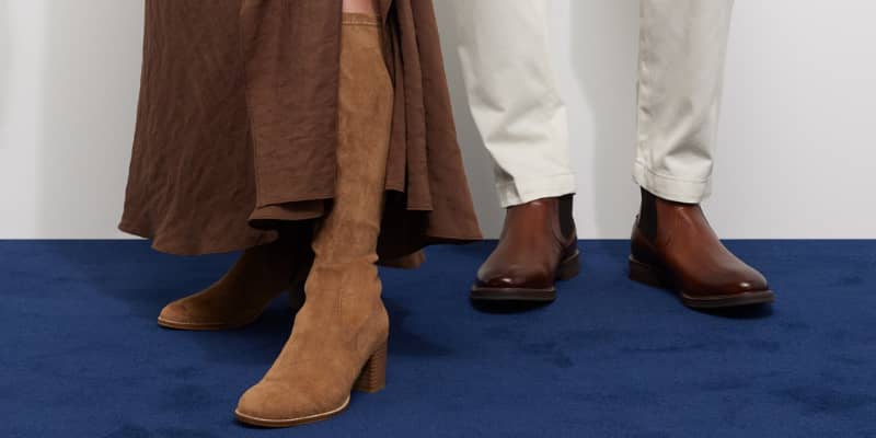 Close-up of a woman's brown suede boots and a man's brown leather Chelsea boots on a navy carpet.