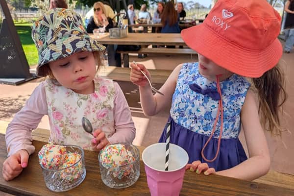 Two young girls in bucket hats sit at a wooden outdoor table, each holding a spoon over a glass dish of ice cream topped with rainbow sprinkles. A pink cup with a striped straw sits between them, with people seated at tables and a lawn in the sunny backgr