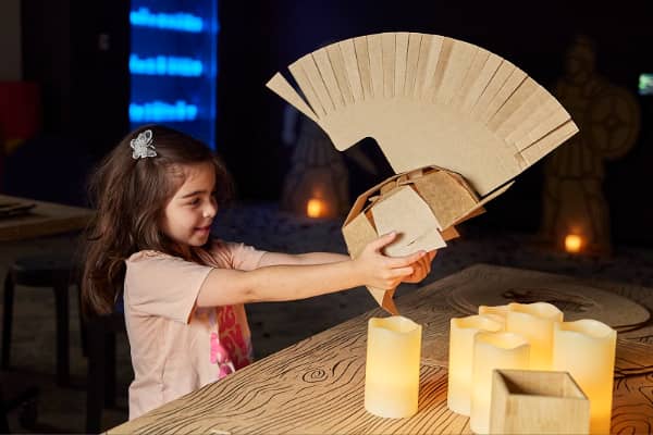 A young girl holds up a cardboard gladiator helmet she has assembled, sitting at a candlelit table inside what appears to be a museum exhibition space inspired by ancient Rome.