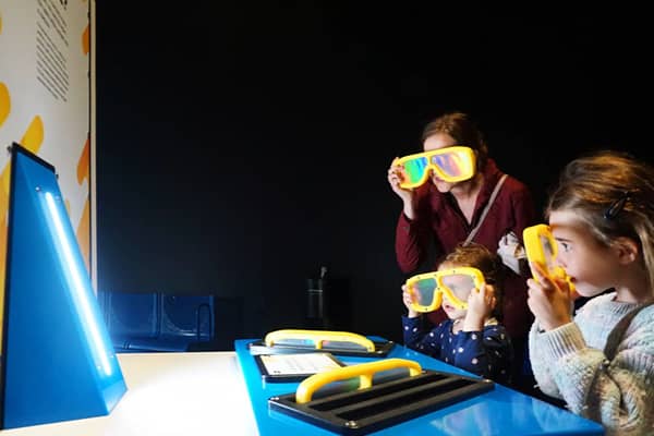 A woman and two young girls look through yellow-framed 3D-style glasses at a glowing colour display, part of an interactive exhibit at Scienceworks. A blue triangular light fixture and clipboards with activity sheets sit on a table in front of them, in a 