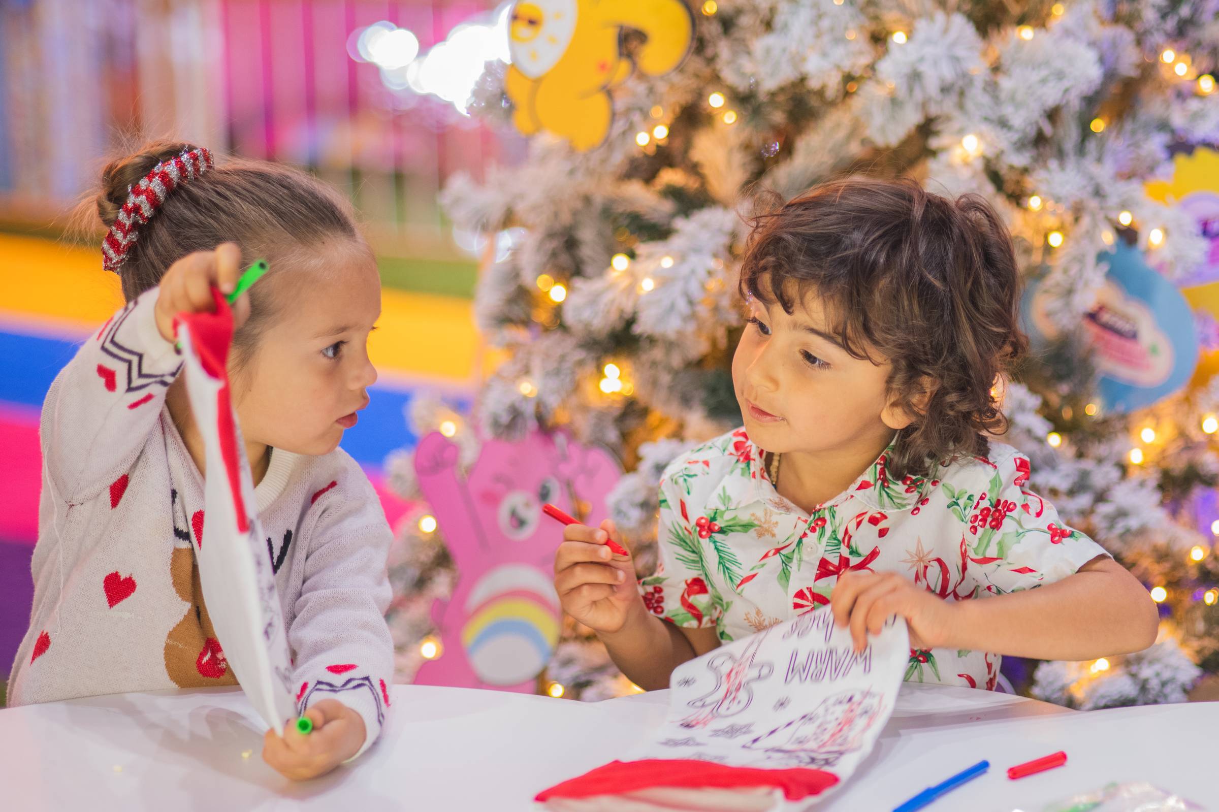 Two young children sit at a white table colouring with markers in front of a white Christmas tree with warm lights. One holds up a red and white fabric stocking and the other draws on a printed sheet, with colourful cartoon bear cutouts in the background.