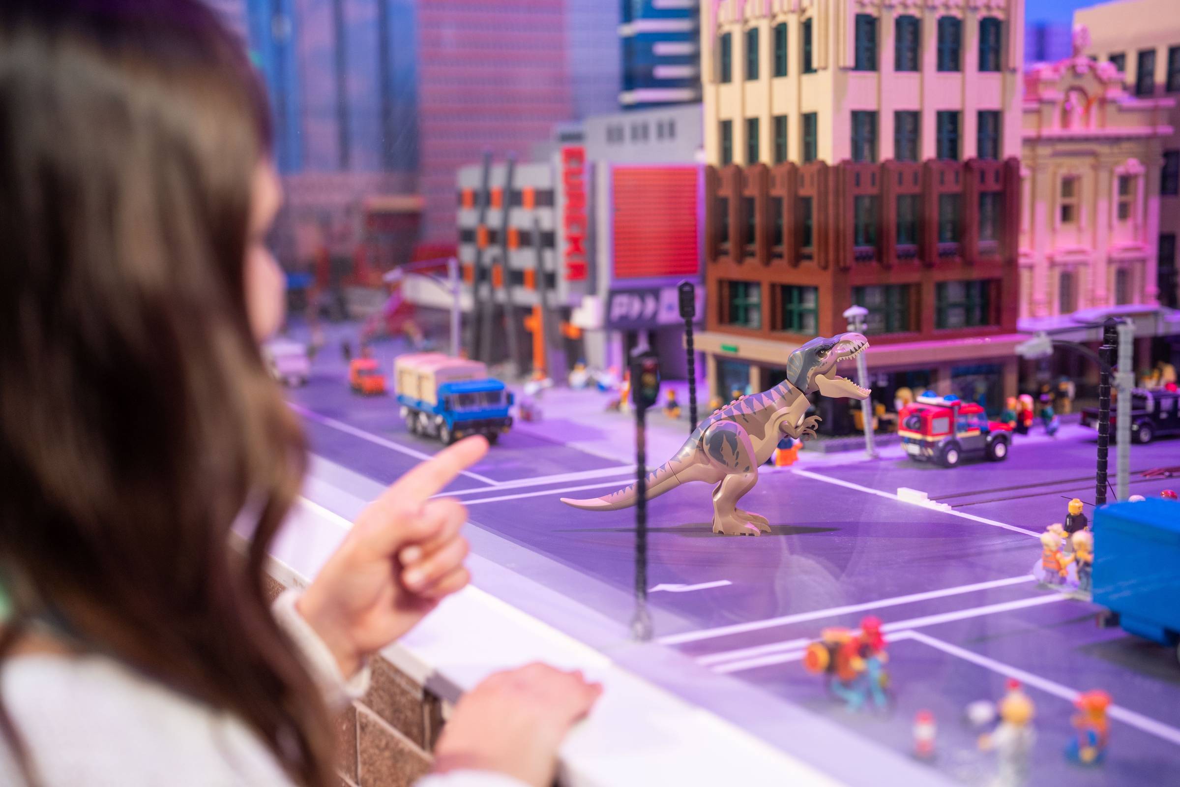A girl points through a glass window at an animated Lego T-Rex standing in a miniature Lego cityscape, with a fire truck, delivery truck and tiny Lego figures nearby.