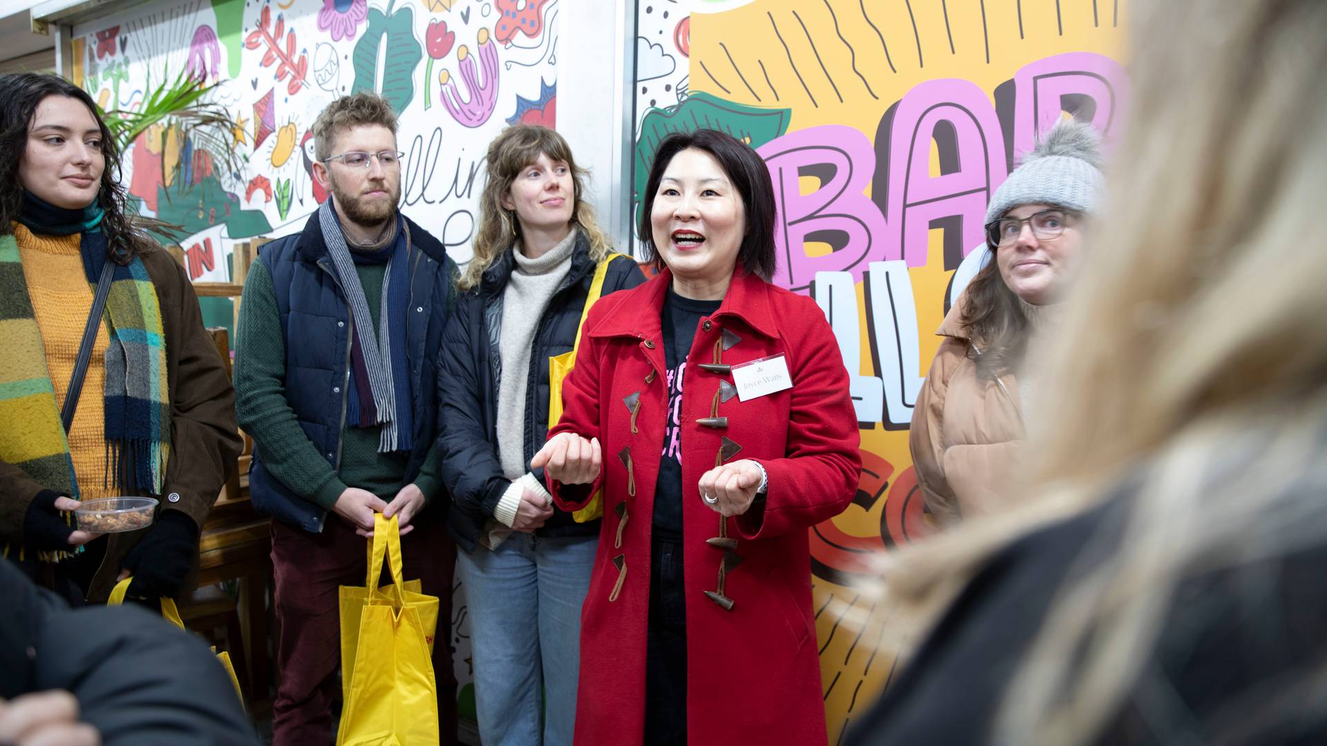 Joyce Watts, in a red coat, talks animatedly to a small tour group holding yellow tote bags inside Footscray Market.