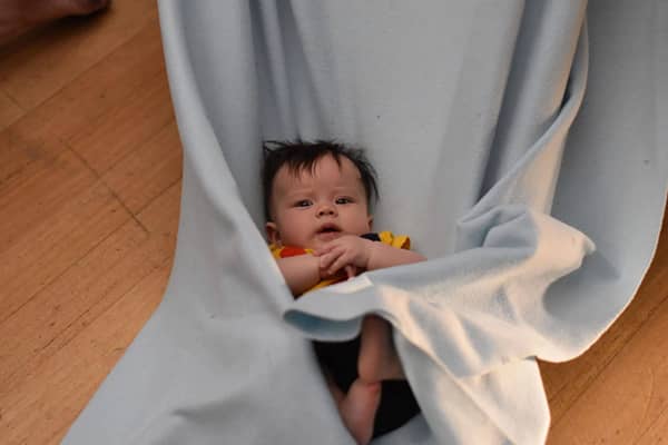 A young baby with dark hair lies cradled in a pale blue fabric baby-wrap sling, held up by an adult's arms, gazing up toward the camera with one hand near their mouth, on a timber floor background.
