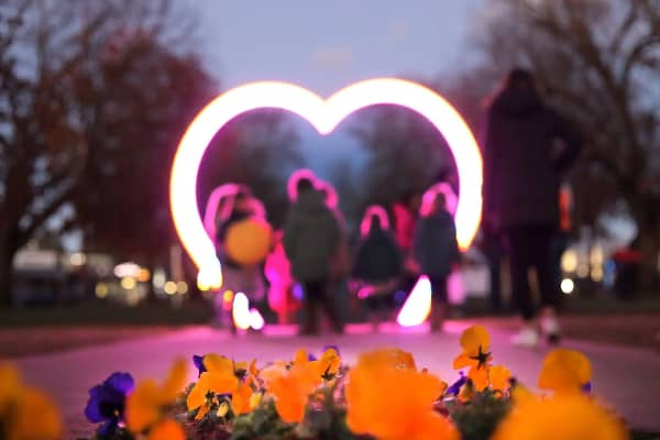 Families gather around a large illuminated heart-shaped light installation at dusk, with orange and purple pansies in the foreground and bare winter trees softly lit in the background.