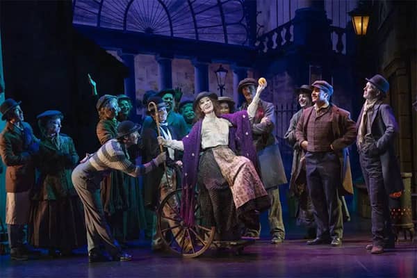 Cast of My Fair Lady on stage, with a woman in a purple shawl and patterned skirt standing on a wooden cart and holding up an orange, surrounded by cast members in worn Edwardian-style street clothes and flat caps, in front of a lamplit street set.