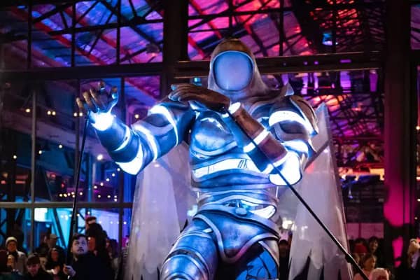 A performer in a dramatic illuminated silver costume with glowing accents takes centre stage at an indoor winter festival event, with a crowd watching under a lit industrial ceiling.