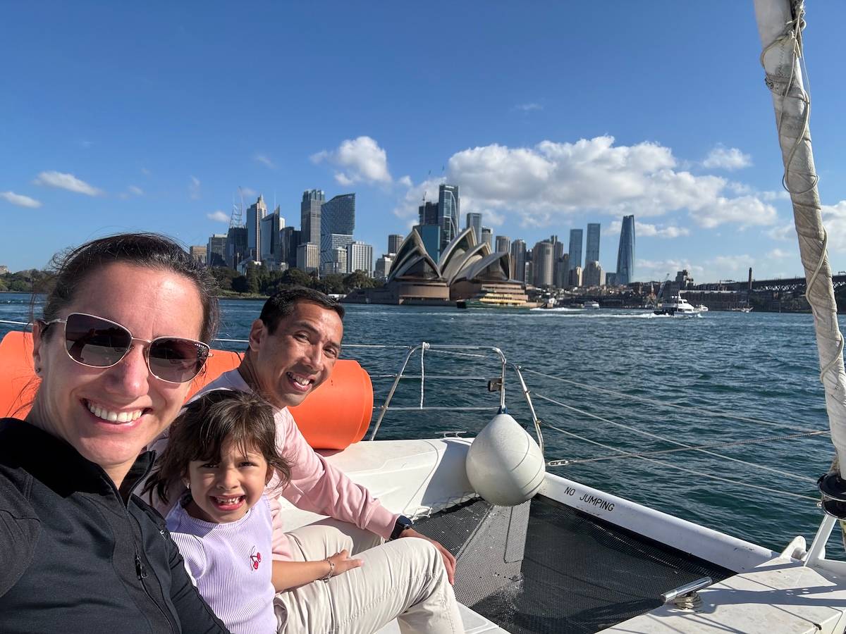 A woman, man and young child smile for a selfie aboard a sailing catamaran on Sydney Harbour, with the Sydney Opera House and CBD skyline visible across the water under a mostly clear blue sky. A red rescue buoy and rigging are visible on the boat's deck, with the words "No Jumping" printed on the hull beside them.