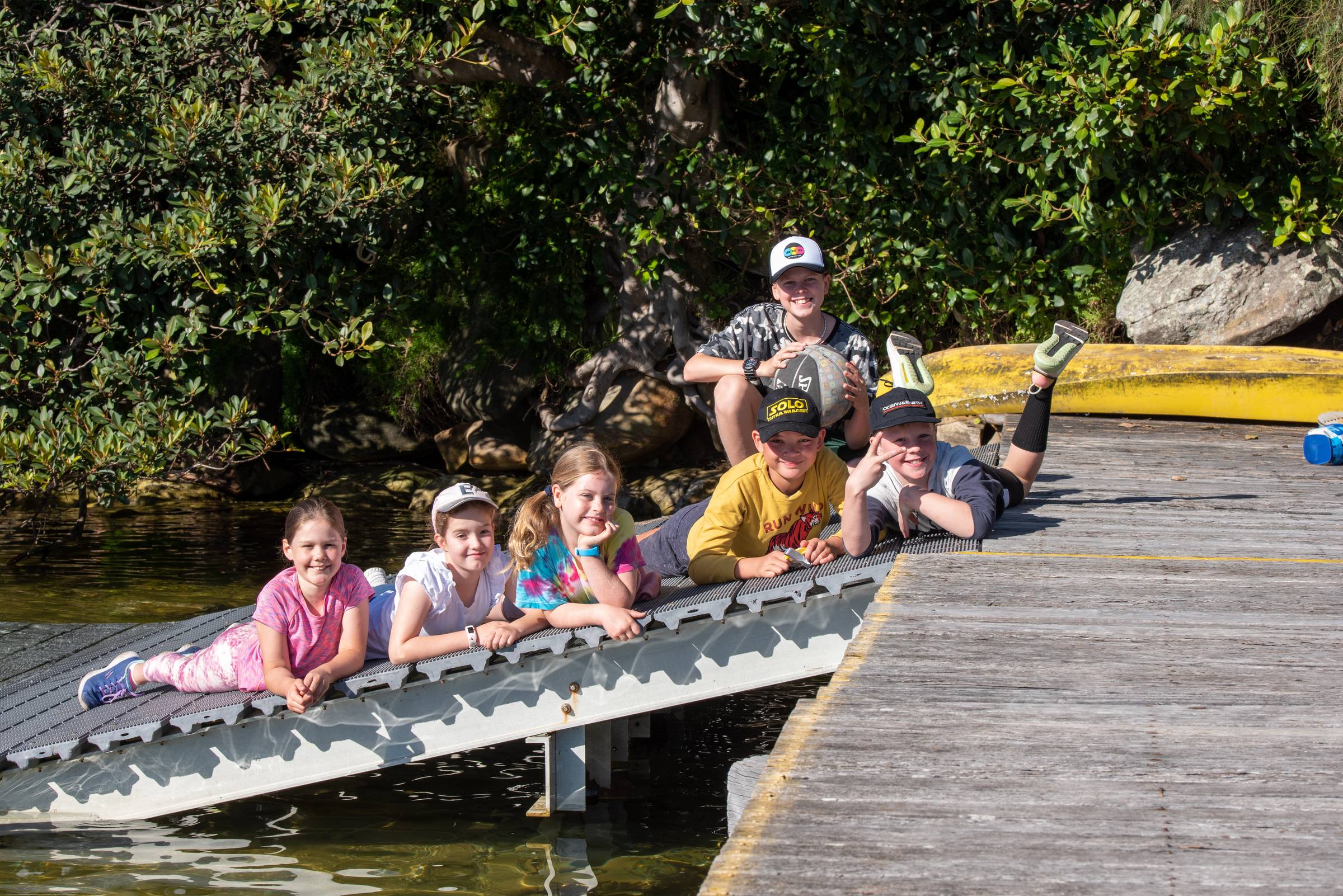 Six children lying and sitting on a wooden jetty over water, smiling at the camera, with a kayak and bushland in the background.