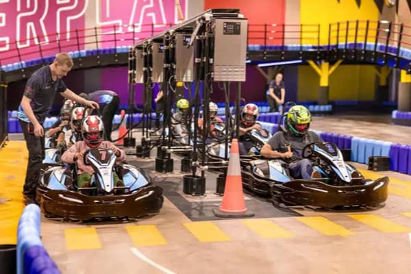 Several young go-karters wearing helmets race on an indoor track, with a staff member assisting one driver at the starting line and an orange traffic cone marking the track edge.