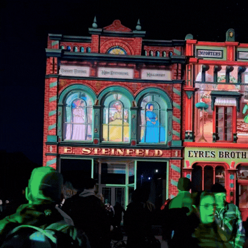 Colourful light projections illuminate historic Main Street shopfronts at Sovereign Hill's Winter Wonderlights, with visitors watching below at night.