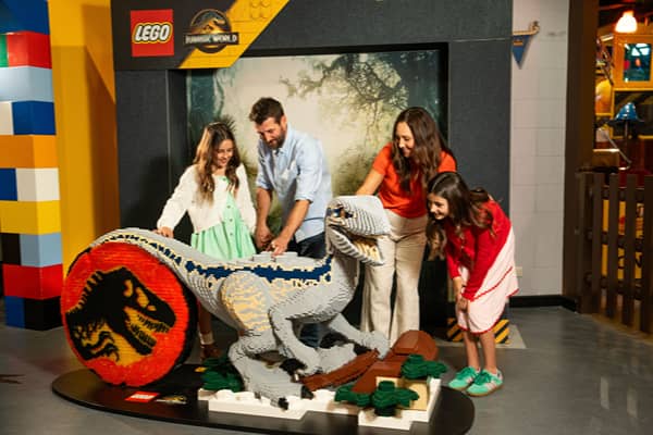 A family of four gathers around a large LEGO brick model of a raptor with blue stripes at LEGOLAND Discovery Centre. A man and two girls lean over to touch the model while a woman in an orange top watches, smiling. Behind them is a Jurassic World LEGO sign and a wall of giant coloured bricks.