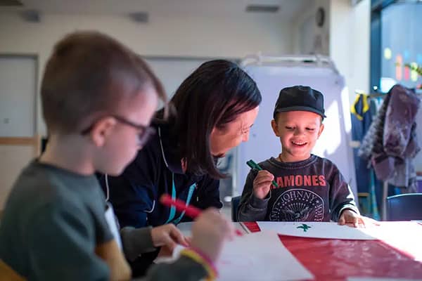 A woman helps two young boys with a craft activity at a table, drawing on red paper with markers. One boy wears glasses and a green jumper; the other wears a black cap and a Ramones-print t-shirt, smiling as he holds a green marker.